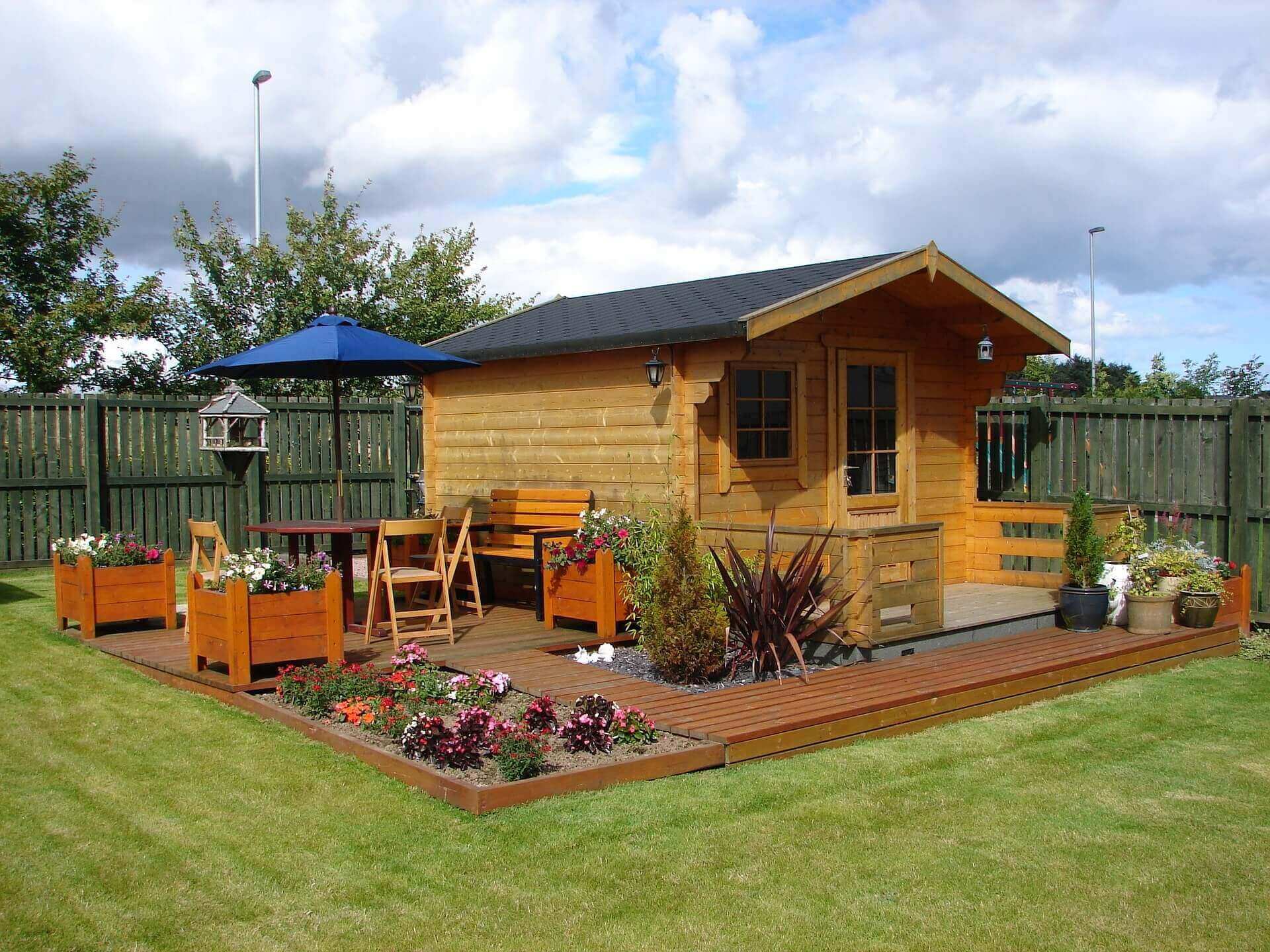 A outdoor shed with attached wooden patio with chairs, a table, and flowers.